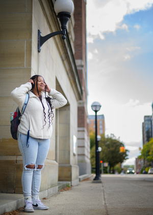 Yessenia Santana-Perez stands on a sidewalk. Her eyes are closed, and she smiles as she listens to music wearing a pair of headphones.