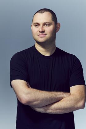 A headshot of Mark Gurman posing with arms crossed against a slate gray background