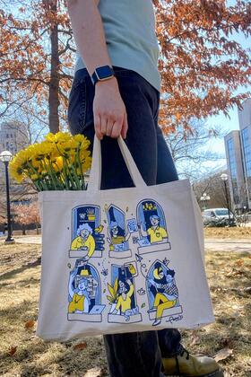 An individual stands outside holding a tote with an illustration, with a bouquet of yellow flowers tucked inside