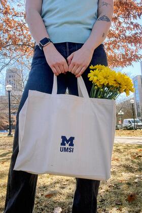 An individual stands outside holding a tote with a blue UMSI logo, with yellow flowers tucked inside