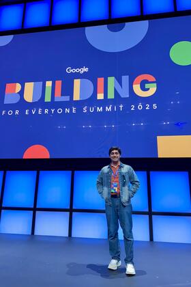 An individual wearing a lanyard smiles on a stage illuminated in blue, with a slide behind them that reads "Google Building for Everyone Summit 2025"