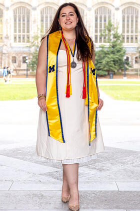 A student wearing a maize stole, red cords, and a medal poses in front of an academic building with tall windows