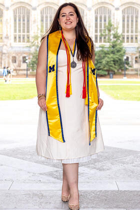 A student wearing a maize stole, red cords, and a medal poses in front of an academic building with tall windows