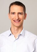 Tomáš Jagelka, a person wearing a light colored striped button up shirt, with short dark hair, smiling. 