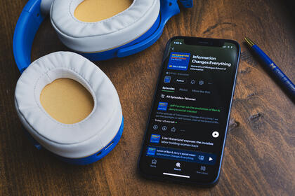Blue and yellow headphones rest on a dark wood background beside a phone with the Information Changes Everything podcast displayed on Spotify. The latest episode is highlighted.