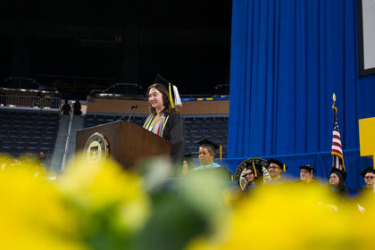 A side view of Charlotte Parent speaking at a podium, with UMSI faculty visible on the stage behind her. In the foreground, there are out-of-focus yellow flowers. 